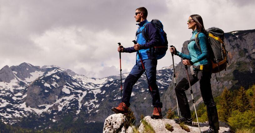 Zwei Wanderer stehen mit Trekkingstöcken auf einem Felsen vor schneebedeckten Bergen